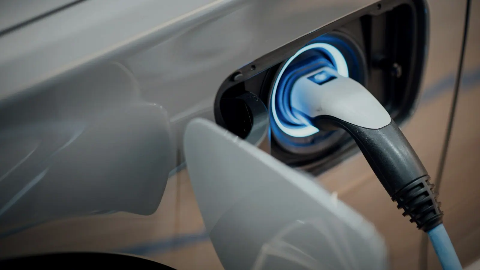 A charging connector plugged into an electric vehicle's charge port, lit blue.