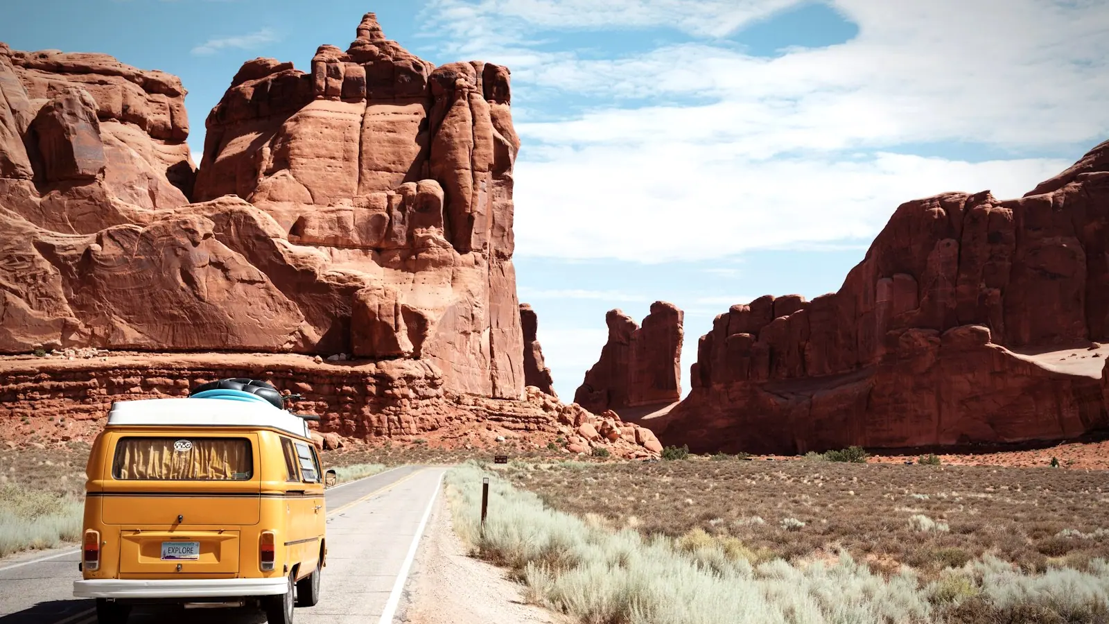 A campervan on an open desert highway winding between red-rock cliffs.