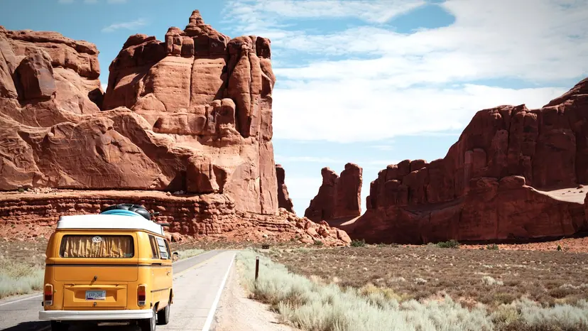 A campervan on an open desert highway winding between red-rock cliffs.