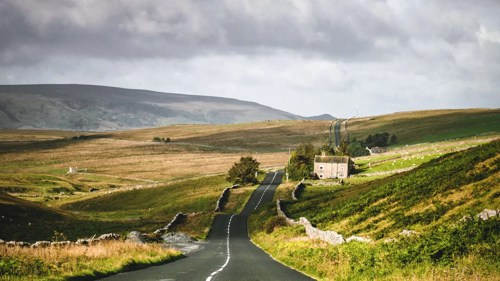 A country road winding through green British hills under a dramatic sky.